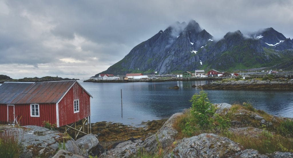 Picturesque Nordic village with wooden huts by a fjord, surrounded by snow-capped mountains.