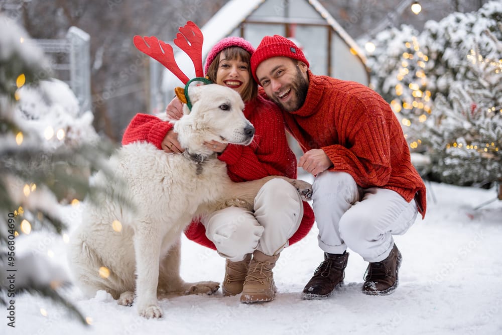 Lovely couple hug with their cute dog wearing toy deers horns at snowy backyard. Young family spending happy winter time together outdoors