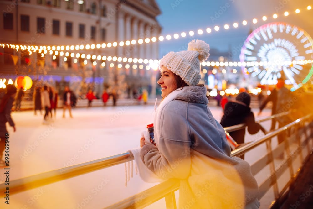 Smiling woman in winter style clothes with coffee near skating rink. Young woman enjoying winter holidays on Christmas market. Lights around.